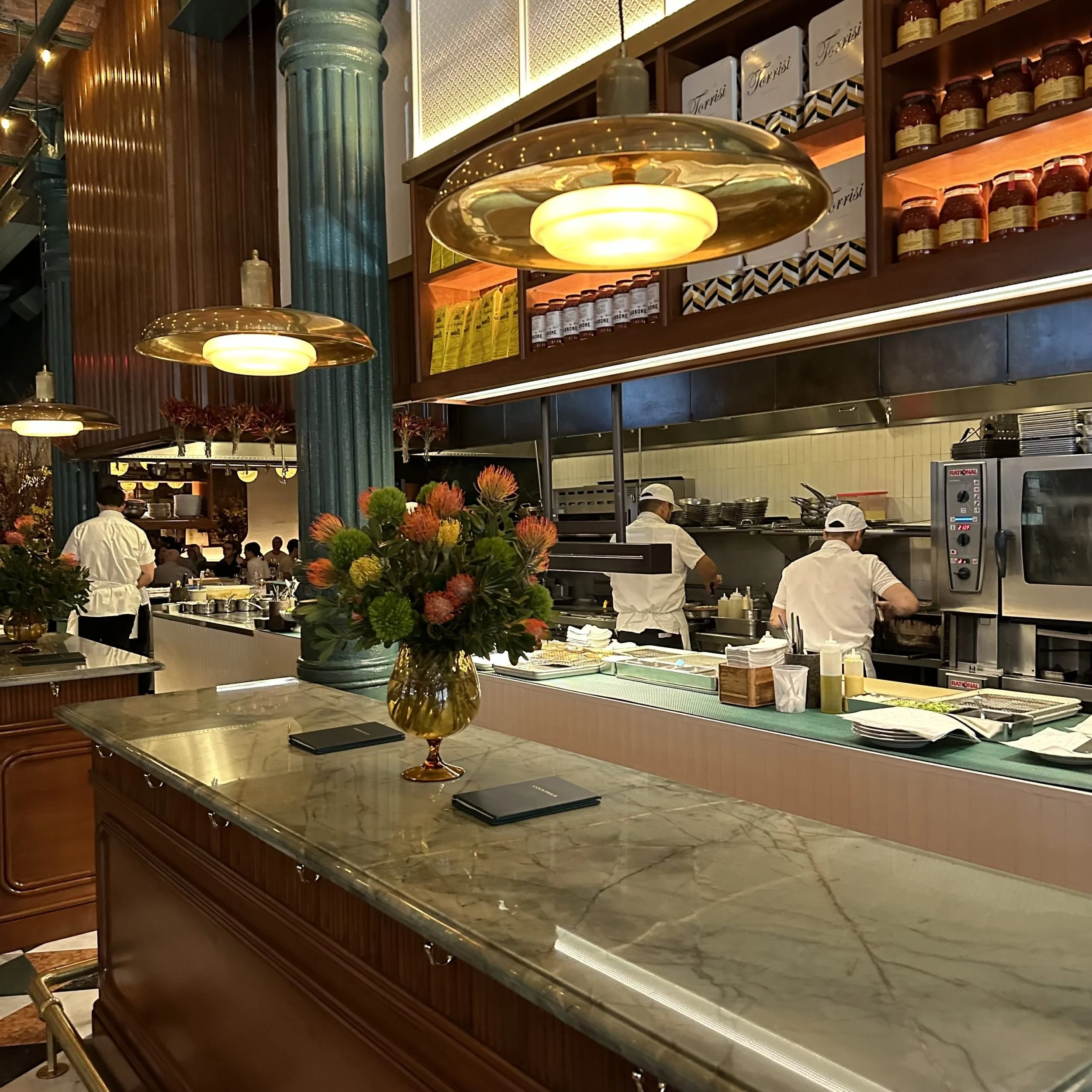 Torrisi in New York City, New York, NY. A restaurant with a marble-topped counter, large ceiling lights, a green column, and shelves stocked with jars and boxed items. Chefs are working behind the counter.