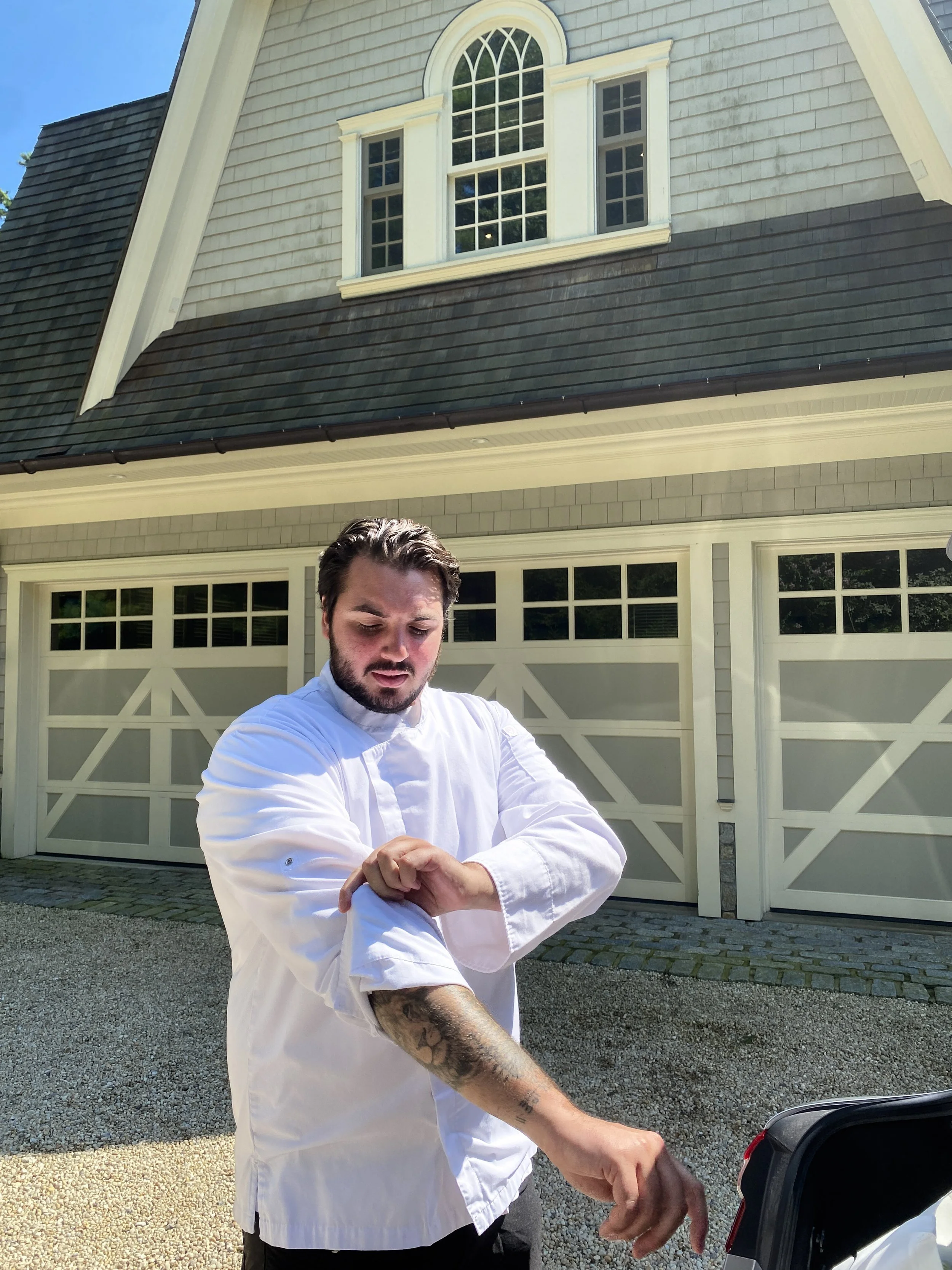 Jonathan Chesler in a white chef's coat standing outside in front of a large house with a garage door. He is looking down and adjusting something with his hands, which are close to a vehicle's trunk.