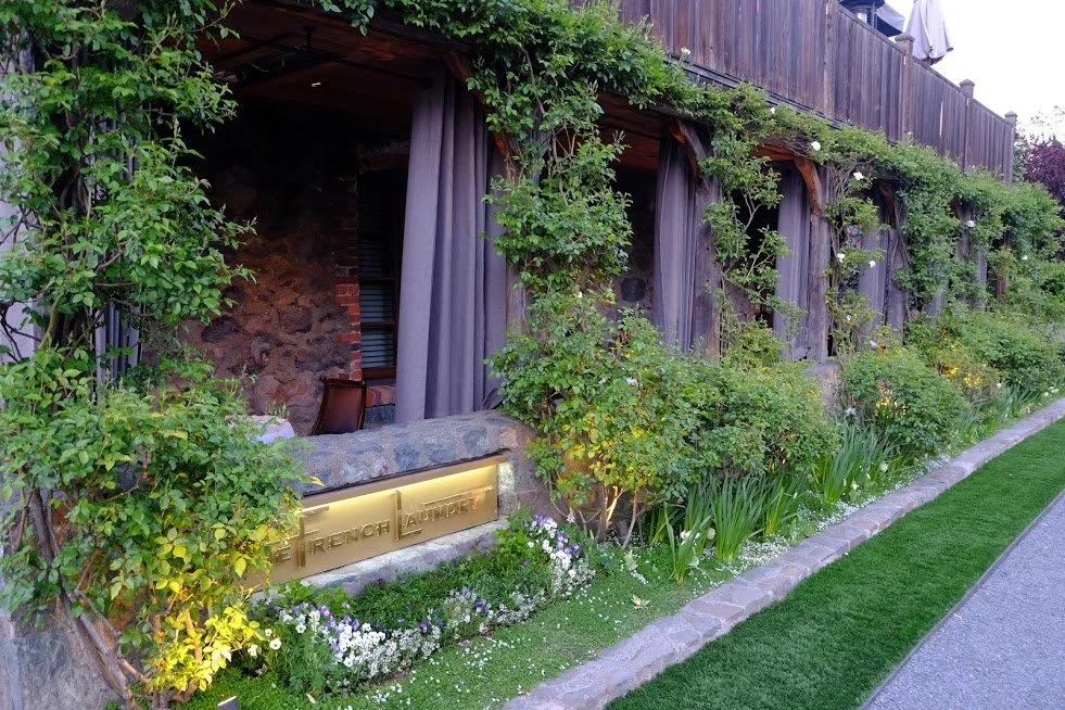 The French Laundry in Yauntville California. Exterior of building wooden fencing and lush greenery, including bushes and flowering plants, along a stone pathway.