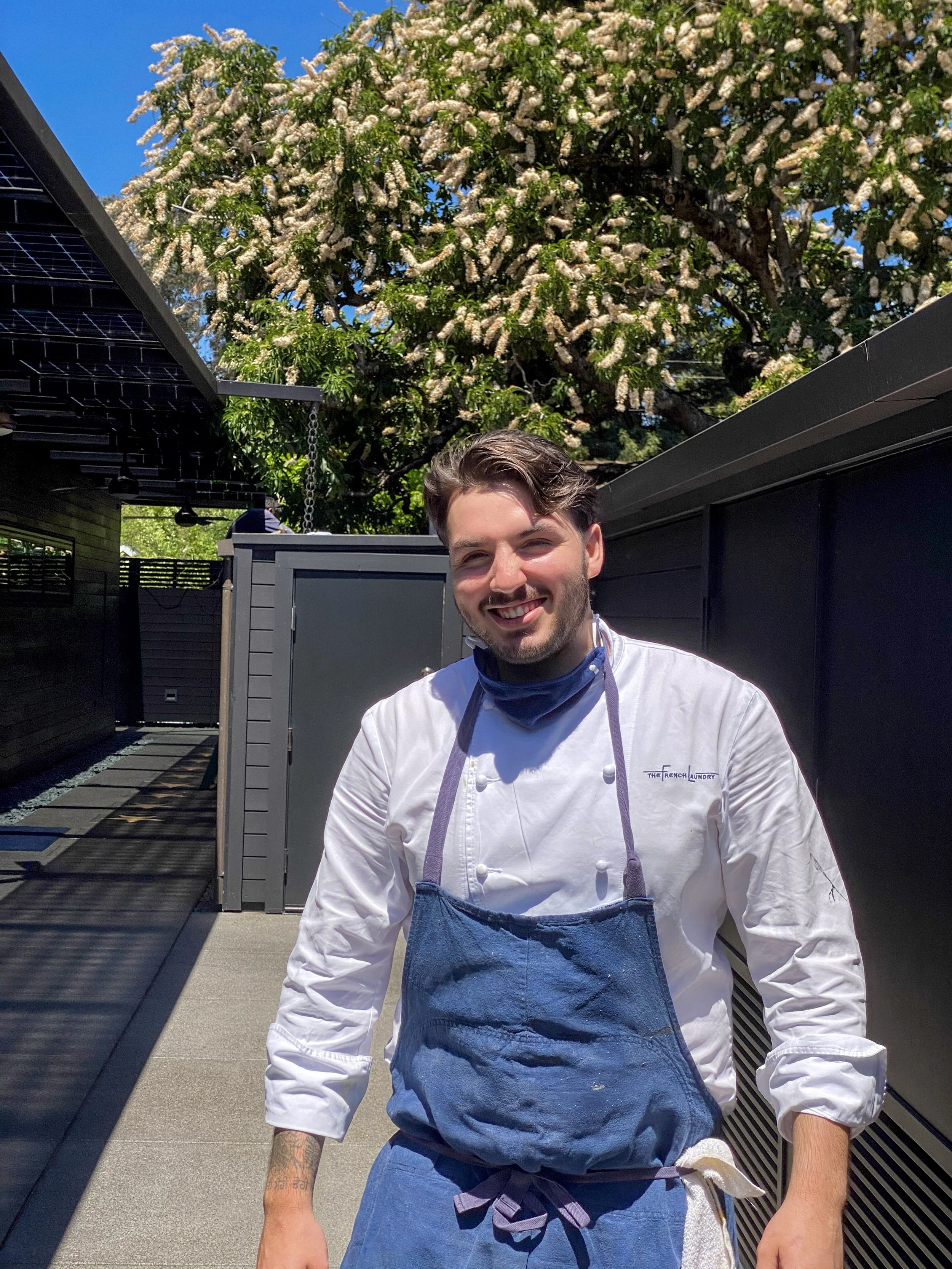 Jonathan Chesler, wearing a white chef's coat and a blue apron, stands outdoors on a sunny day. Behind him is a large flowering tree with white blossoms and a clear blue sky.