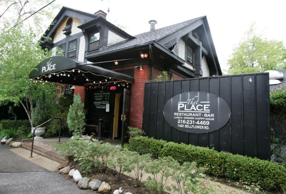 Exterior of a restaurant and bar called 'That Place,' with a black awning decorated with string lights, situated in a building with dark gray and brick walls, surrounded by greenery and rocks.