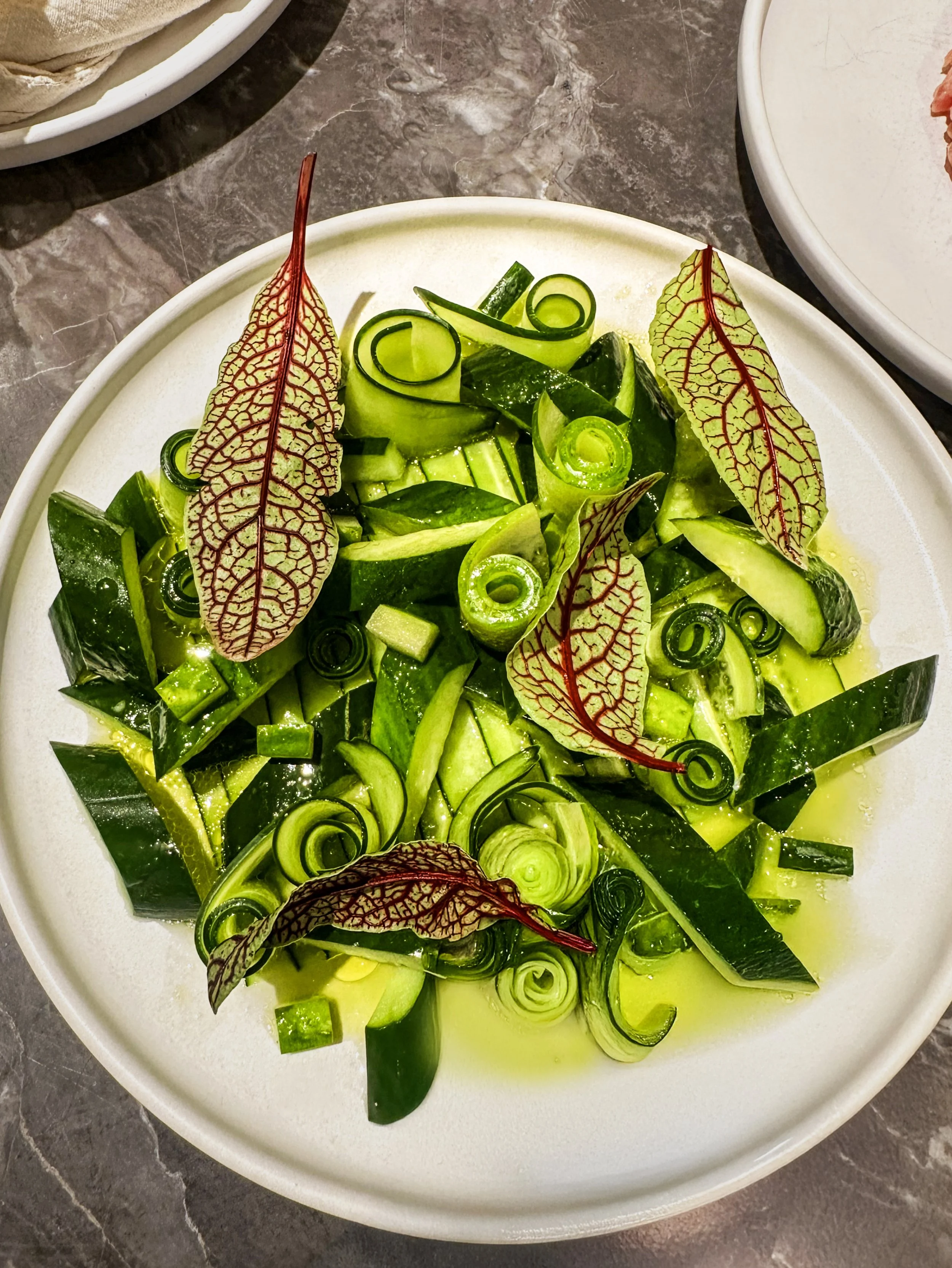 A white bowl filled with sliced cucumbers and garnished with red-veined beetle leaves, on a dark marble surface.