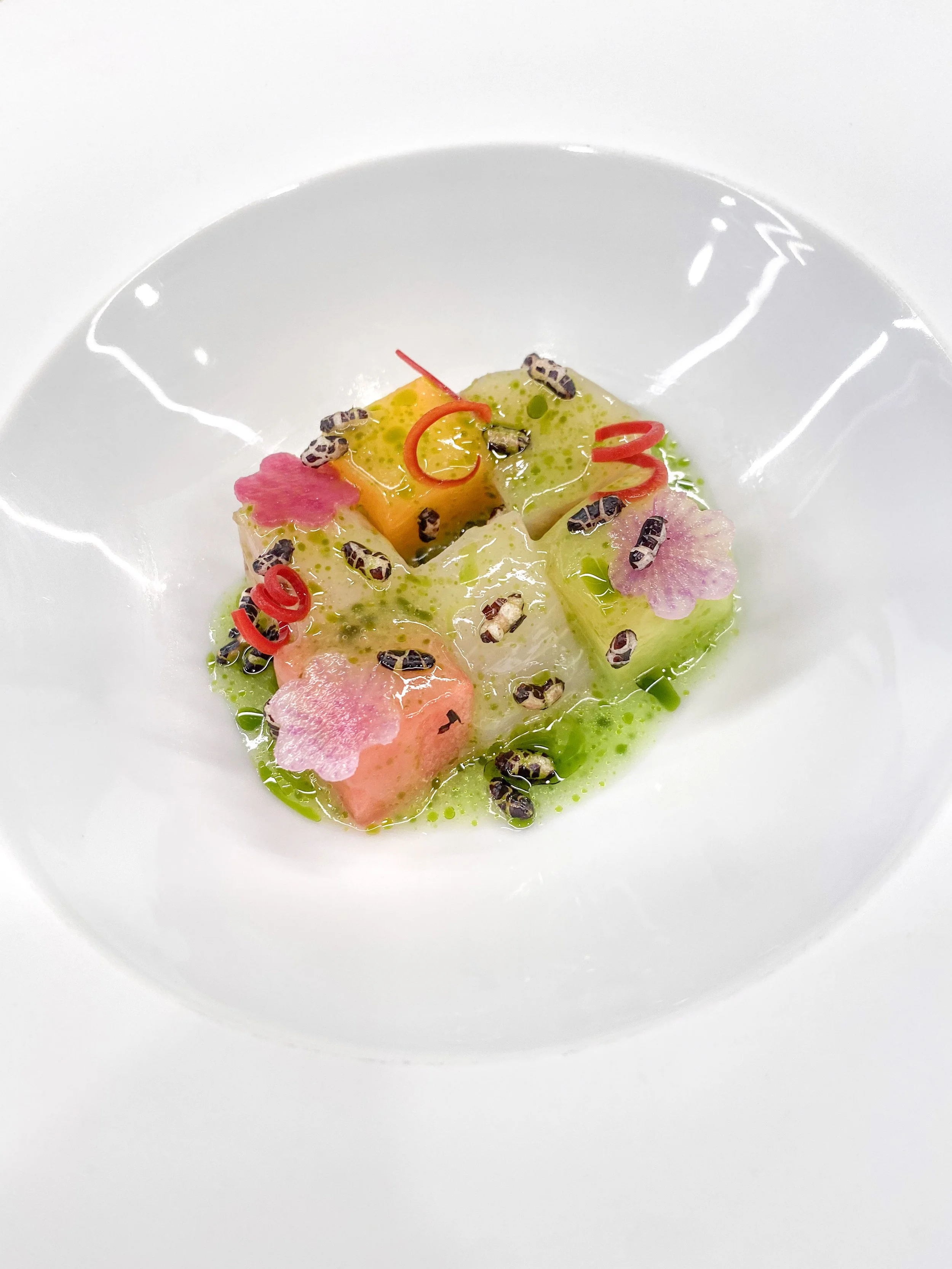 A white bowl containing a colorful gourmet dish with pink, yellow, and green edible flower slices, microgreens, and black seeds, garnished with red pepper curl and oil drizzle.