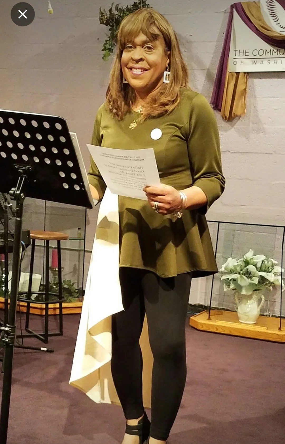 A woman standing indoors, smiling, holding a paper, wearing a green top, earrings, and rings, with a music stand and decorative items in the background.