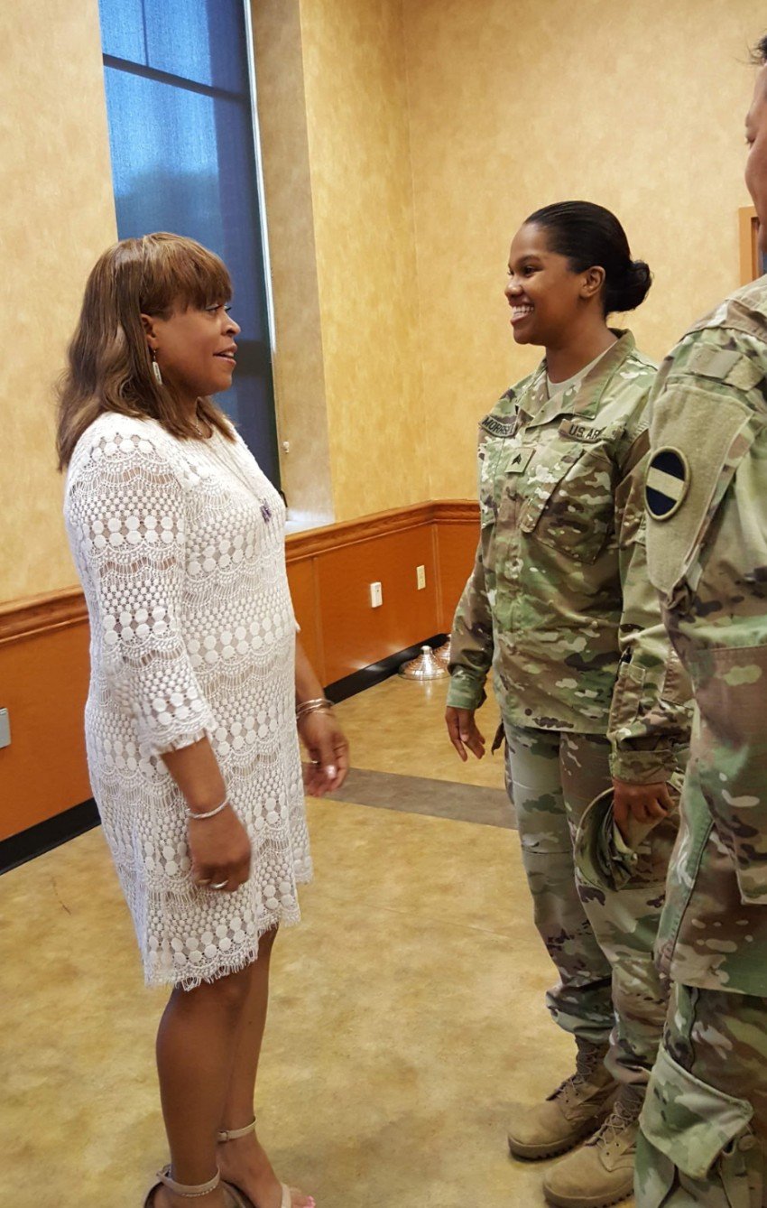 A woman in a white crochet dress talking to two female soldiers in camouflage uniforms inside a room with yellow walls and a wooden bottom panel.