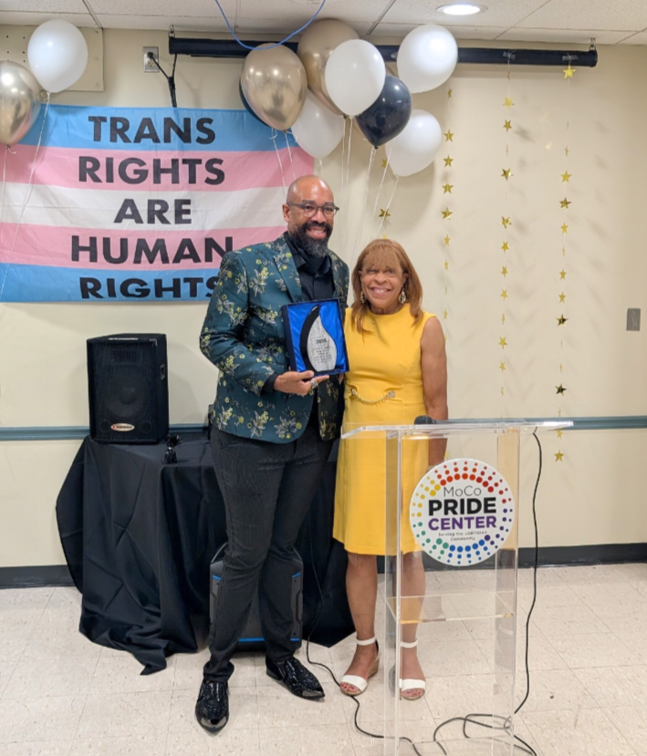 Two people, a man and a woman, standing together at an event. The man is holding an award. Behind them is a banner that reads 'Trans Rights Are Human Rights' with the transgender pride flag colors and a cluster of balloons. To their right, a clear podium has a logo that says 'MoCo Pride Center,' and the background has gold star decorations.