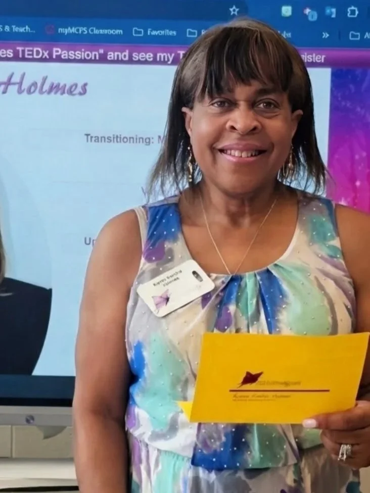 A woman smiling and holding a yellow envelope, wearing a colorful galaxy-patterned dress with a name tag, standing in front of a presentation screen.