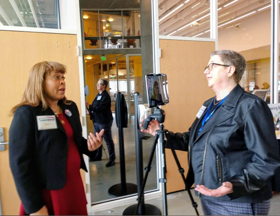 A woman being interviewed or giving a statement to a woman with short gray hair and glasses, using a camera on a tripod in a modern indoor setting.