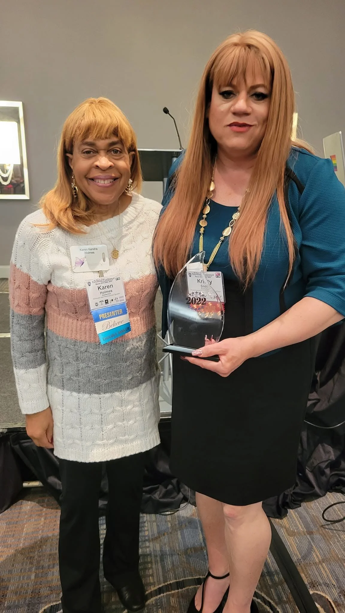 Two women standing together at an event, one is holding an award; both are smiling and wearing name tags.