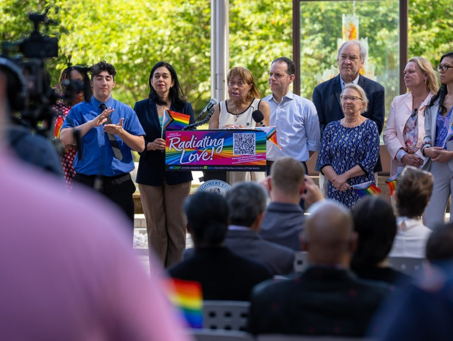 People gathered at an outdoor LGBTQ+ Pride event, including a speaker at a podium with a rainbow banner, surrounded by individuals holding rainbow flags and wearing bright clothing, with onlookers in the foreground and lush greenery in the background