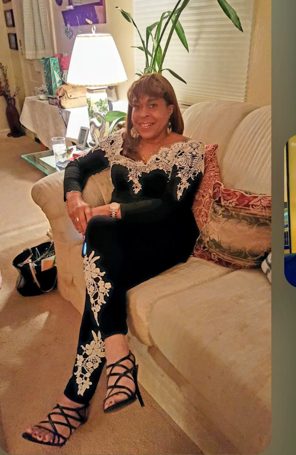 A woman sitting on a beige couch in a living room, wearing a black dress with intricate gold embroidery, black high heels, and jewelry, smiling at the camera. The room has a lamp, plants, and decorative pillows, with a side table holding a glass of w