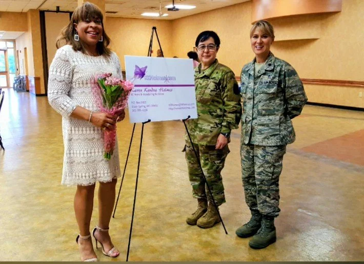 Woman in a white lace dress holding a bouquet of pink flowers, standing next to two women in military camouflage uniforms, in a large indoor room with a sign on a stand.