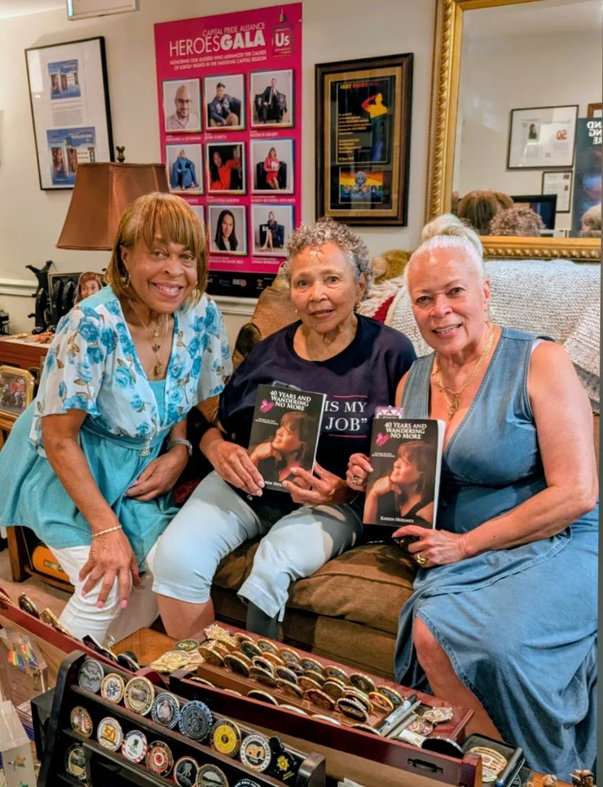 Three women sitting on a couch at a book signing event, holding books, surrounded by framed pictures and posters on the wall.