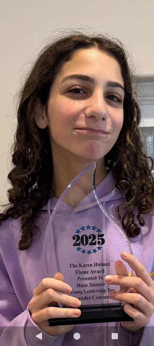 Young girl with curly hair holds a glass award with the year 2025 engraved on it, wearing a pastel purple hoodie.