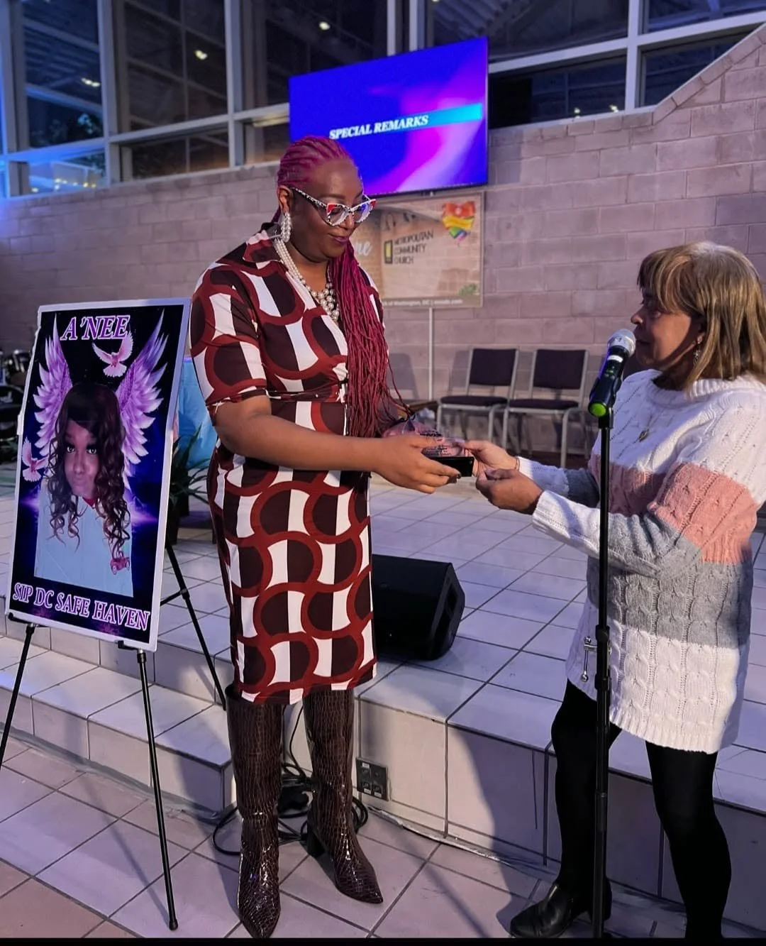 A woman wearing a patterned dress and shiny boots receives an award or a certificate from another woman with a microphone, in an indoor event space. There is a poster with an image of a woman and the words 'A NEE Sip DC Safe Haven' beside them, and a screen in the background displaying 'Special Remarks.'