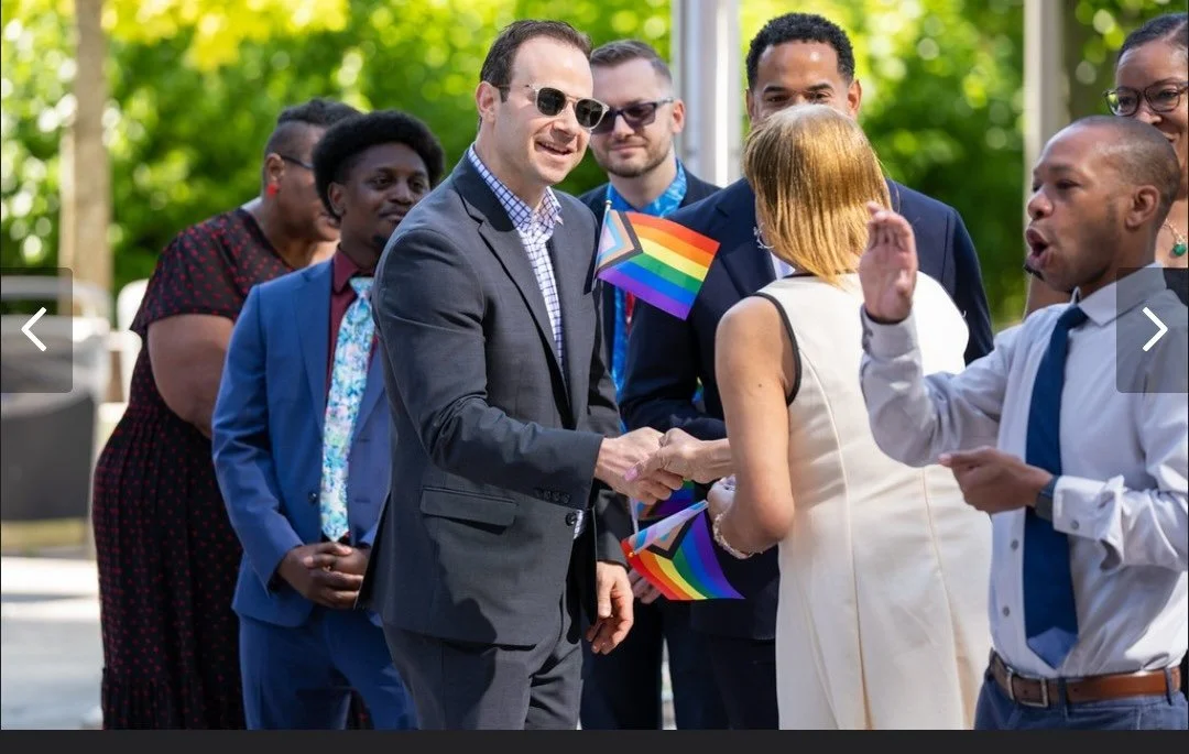 A diverse group of people at an outdoor pride event, with some holding rainbow pride flags. Two individuals are shaking hands while others smile and talk.