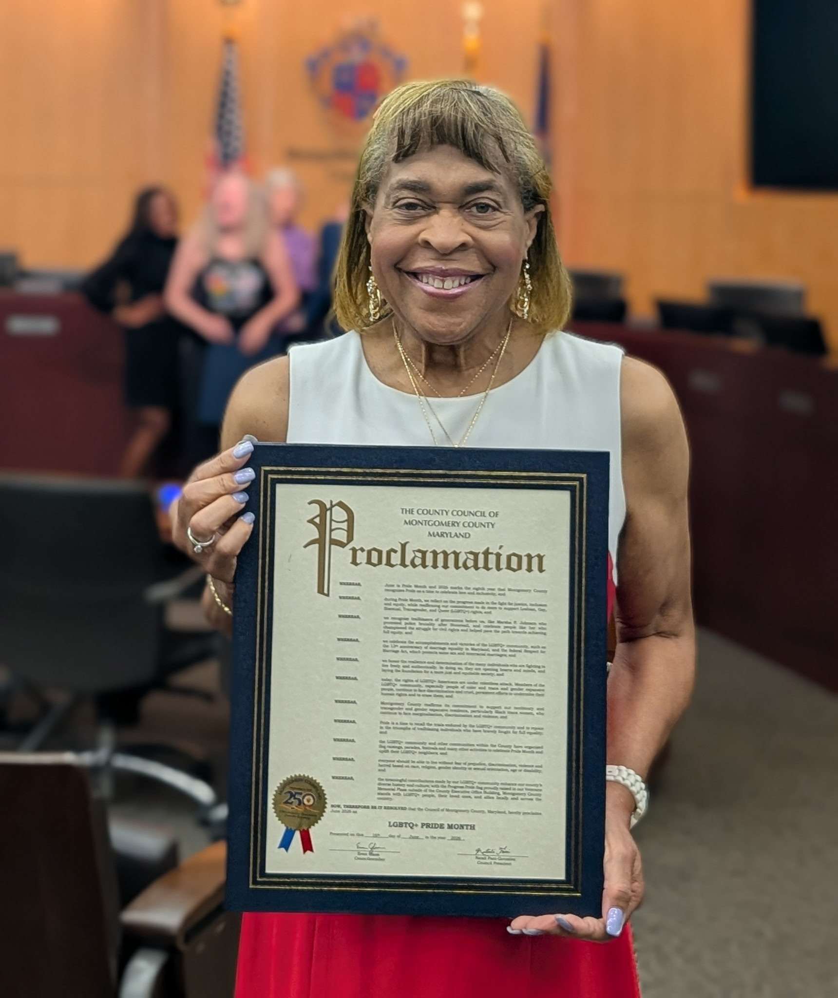 A woman holding a proclamation certificate at an indoor event. The woman is smiling and wearing jewelry, with a blurred background featuring an American flag and a coat of arms.