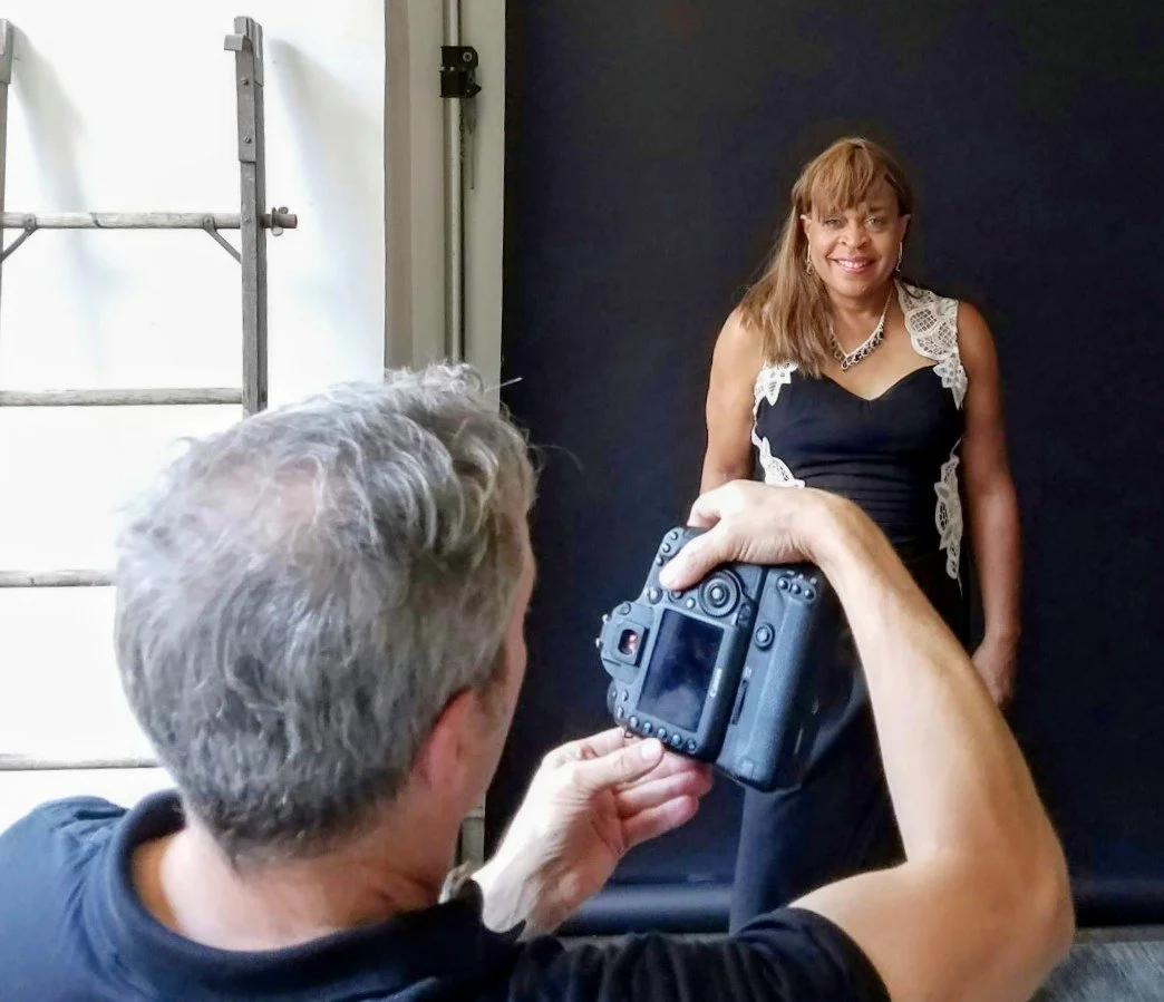 A woman in a black dress with lace details is posing for a photo against a black backdrop while a person takes her picture with a digital camera in an indoor setting.
