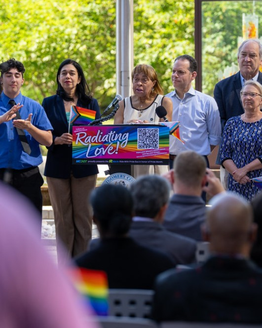 Group of people standing in front of a podium with a sign that says "Radiating Love!" and rainbow flags, during a pride event or rally, with an audience in front.