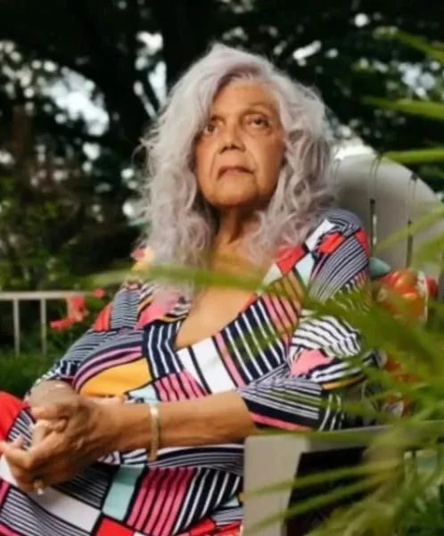 An elderly woman with curly gray hair sitting outdoors in a garden, wearing a colorful striped dress and looking upwards.