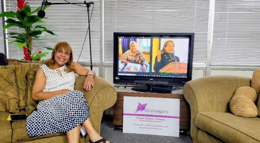 A woman with light brown hair and a polka dot dress sitting on a beige couch, smiling, in an indoor setting with a TV screen on a wooden stand behind her. The TV screen displays two women speaking; the setting appears to be an interview or presentati