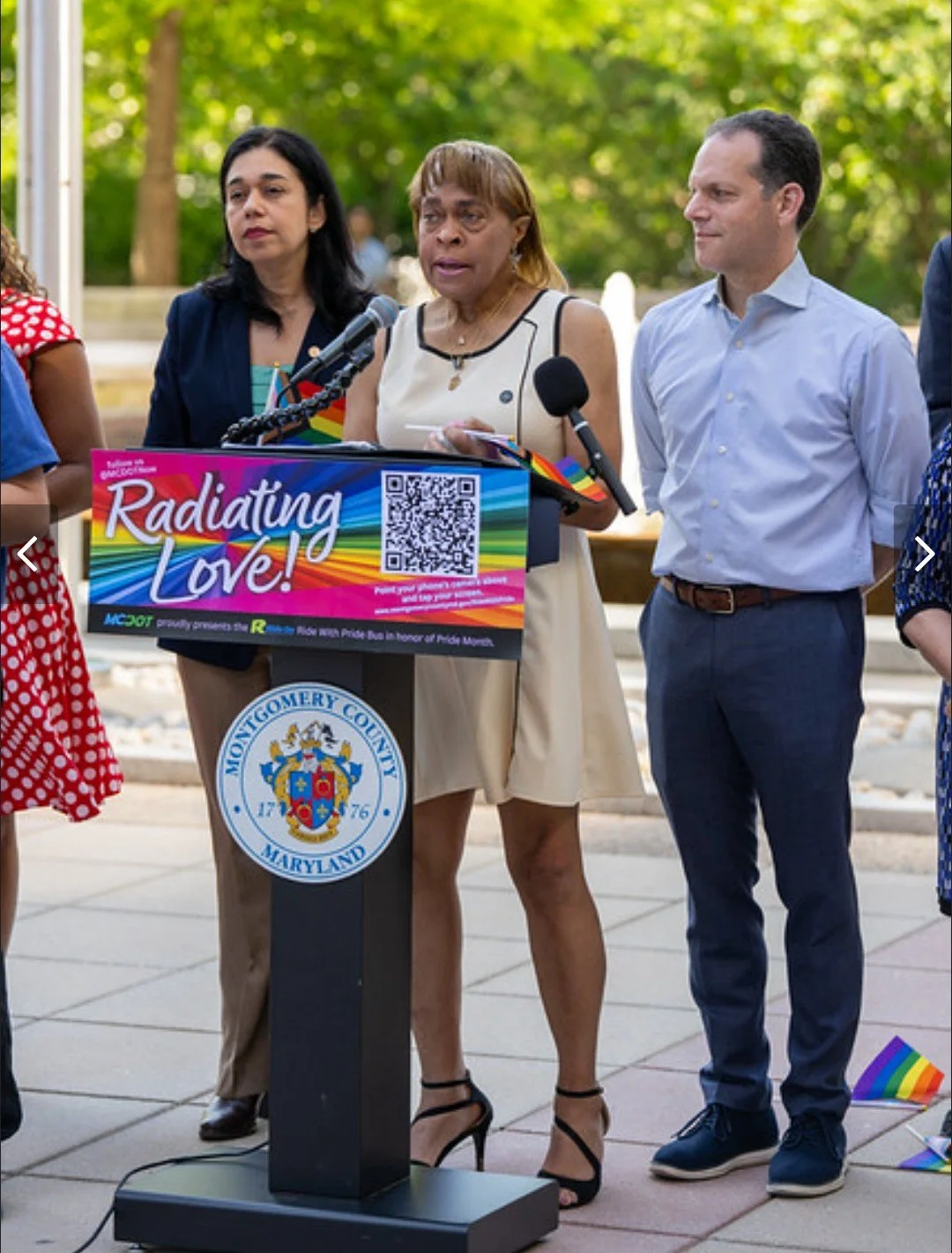 Woman speaking at a podium with a sign that reads "Radiating Love!" at an event for Pride Month, with other people standing behind her outdoors.