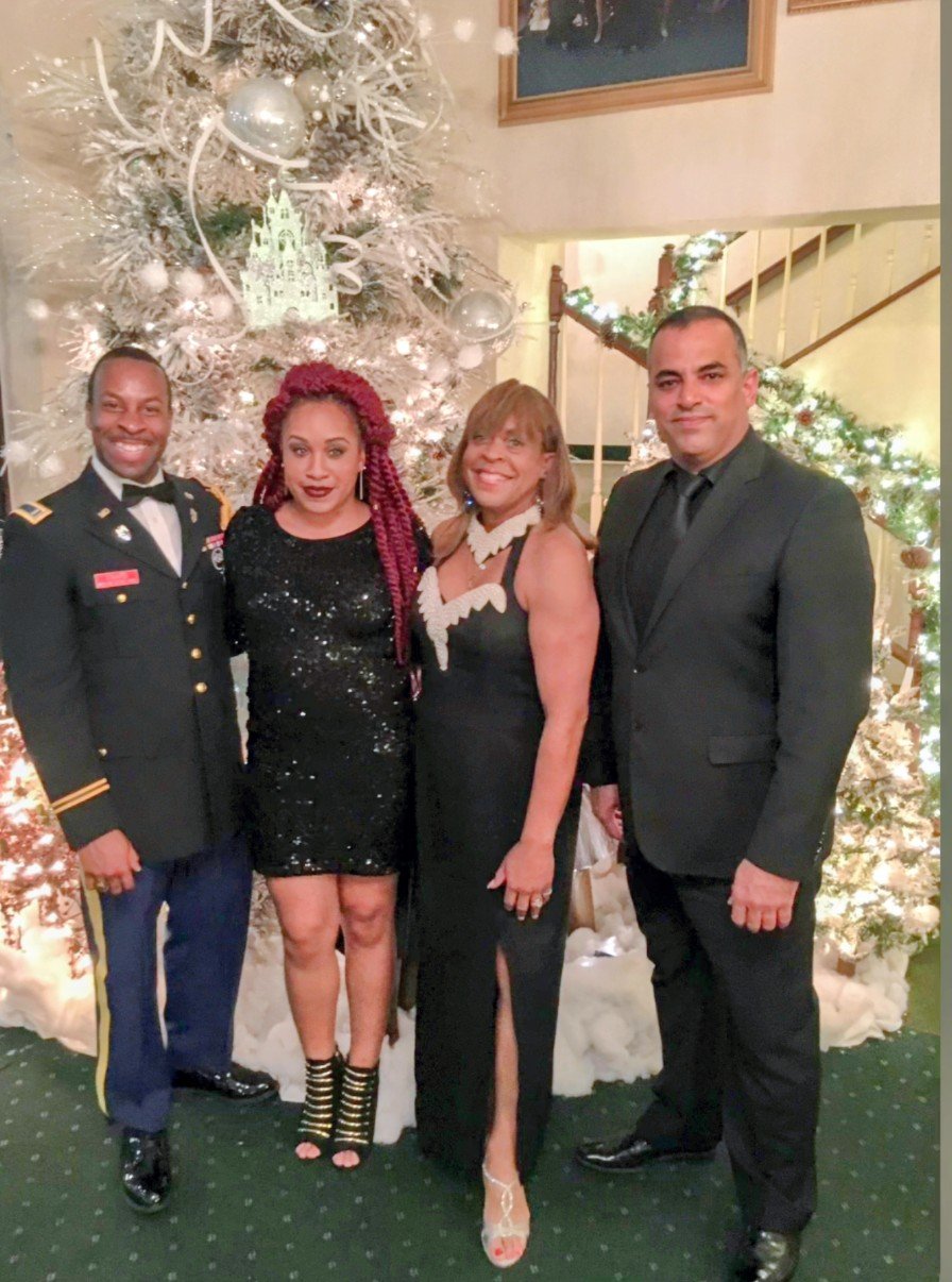 Four people dressed in formal attire standing in front of a decorated Christmas tree with white ornaments, ribbon, and lights in a festive indoor setting.