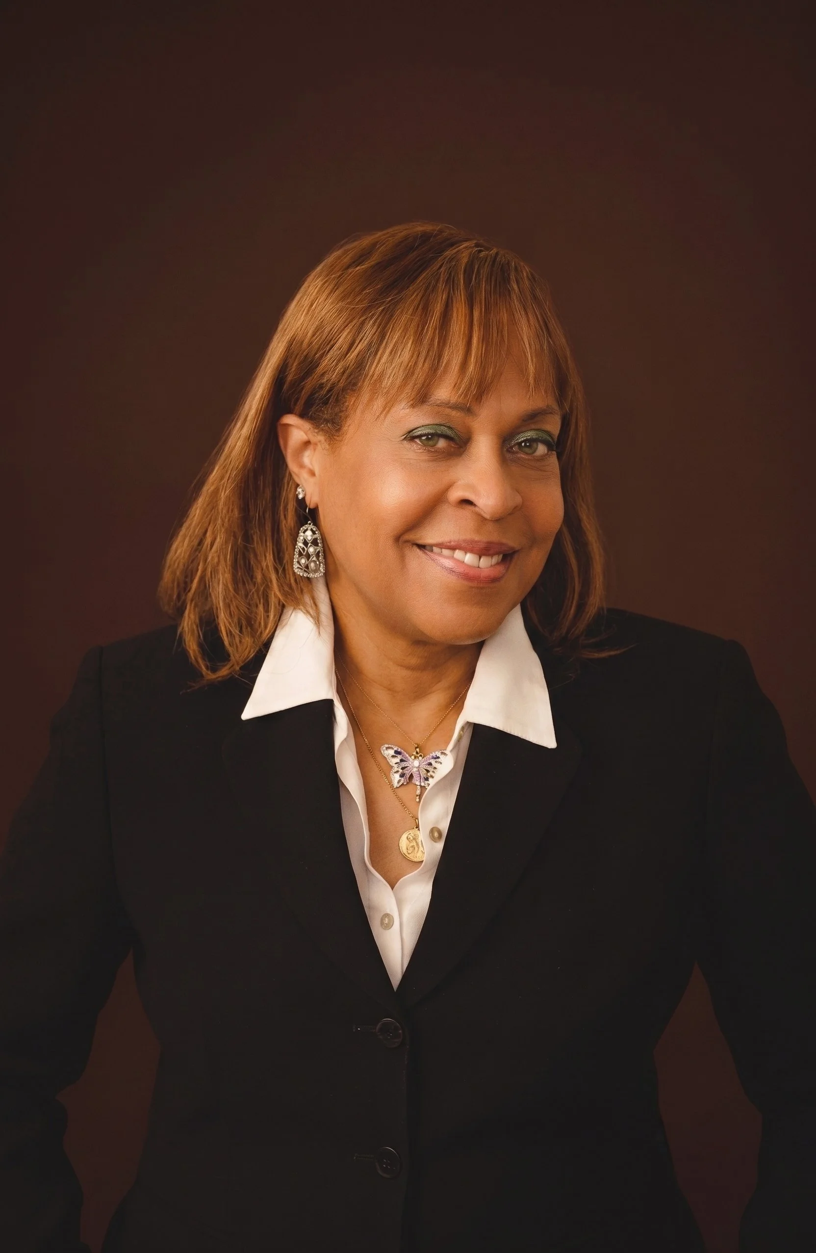 Portrait of a middle-aged woman with light skin, shoulder-length reddish-brown hair, wearing a black blazer, white collared shirt, butterfly pendant necklace, and dangling earrings, smiling against a dark background.