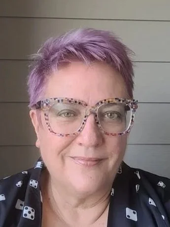 A woman with short, lavender-colored hair wearing large, patterned glasses and a black shirt with a white dice pattern standing in front of a wooden wall.