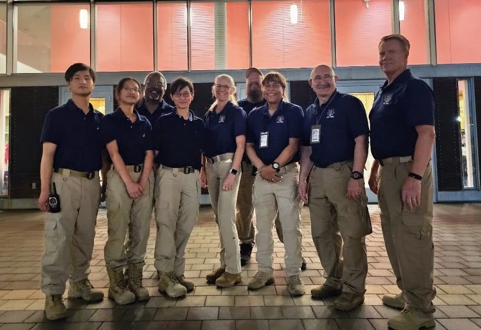 A group of nine people wearing matching navy blue shirts with logos and beige cargo pants, standing together indoors in a spacious area with a brick floor and large windows, smiling at the camera.