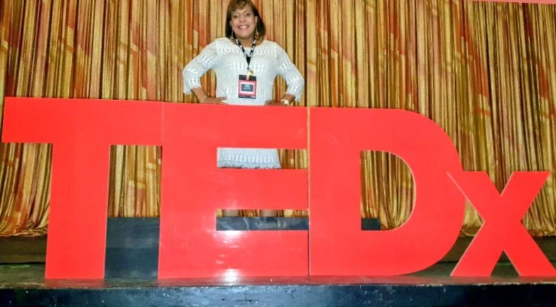 A woman standing behind large red 'TEDx' letters on a stage, with a gold curtain in the background.