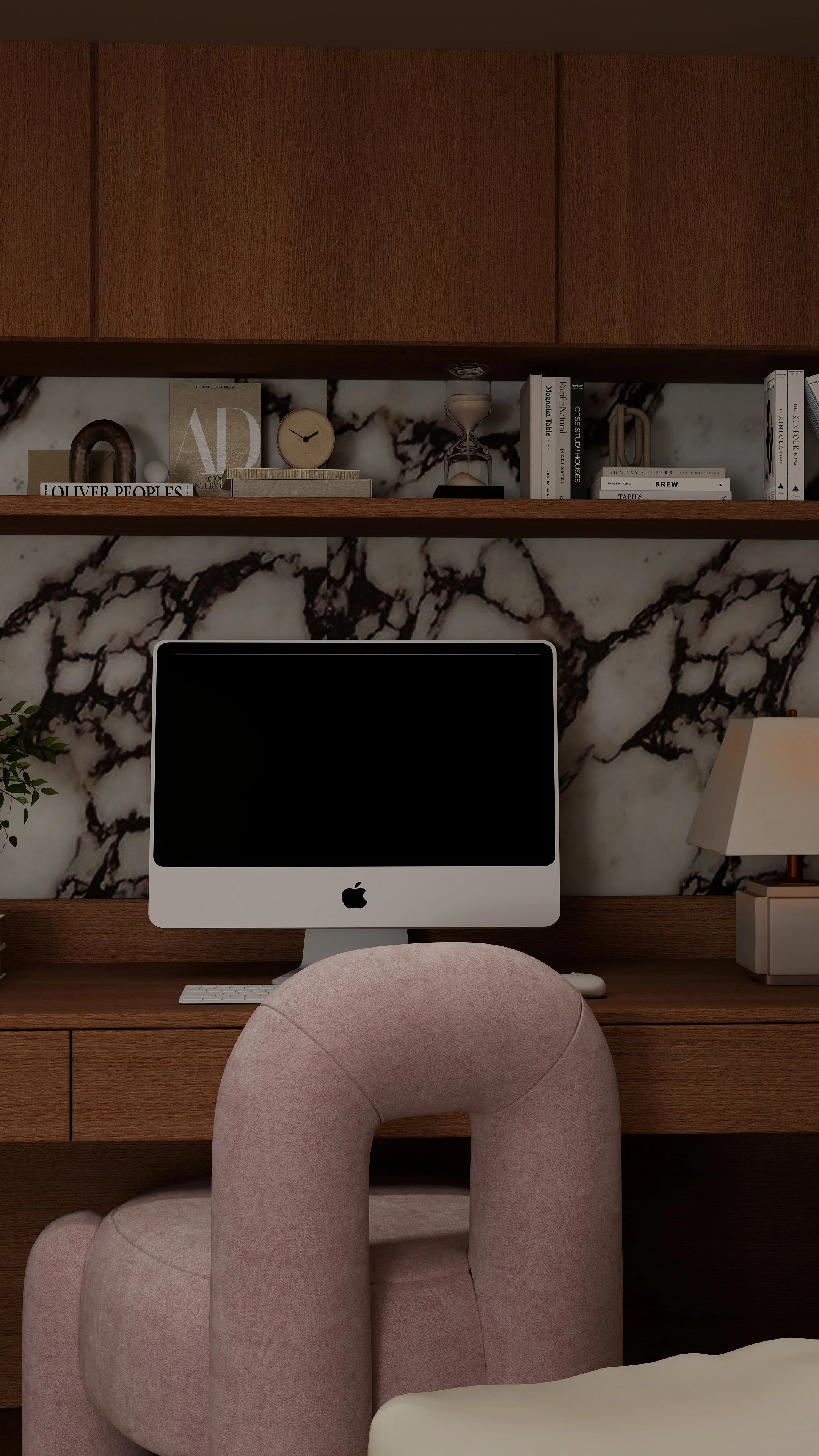 A home office workspace with a pink plush chair, a wooden desk with an iMac computer, a marble wall background, and a wooden shelf holding books, a clock, and decorative items.