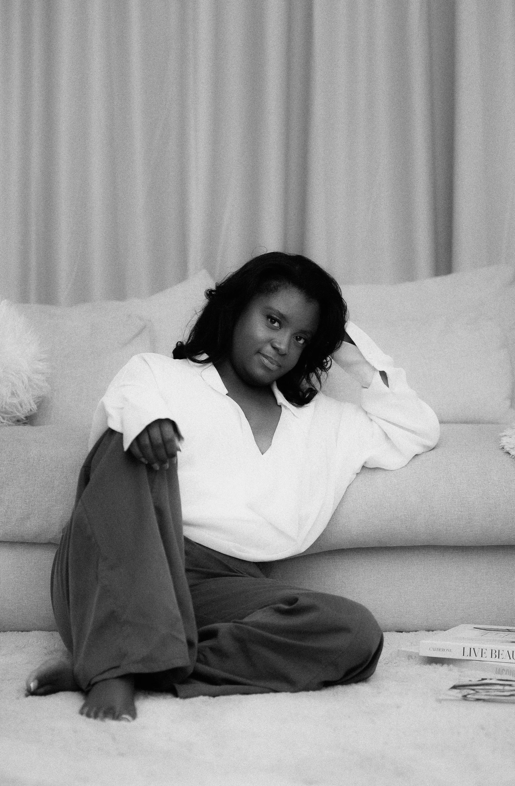 A woman with dark hair sitting on a couch with a relaxed pose, resting her head on her right hand, wearing a white shirt and dark pants, in a room with a curtain background and a stack of magazines on the floor.