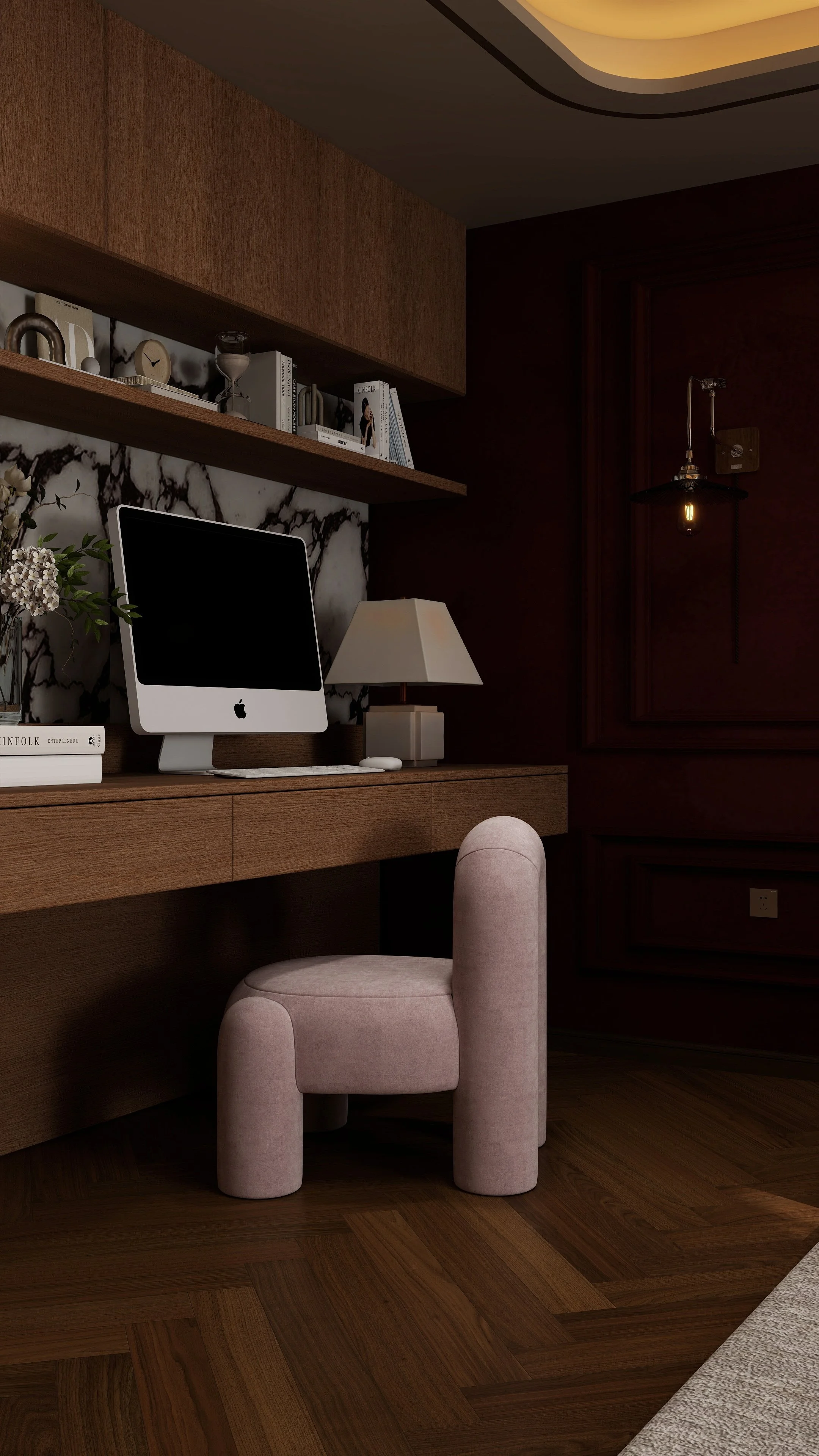 A modern home office with a pink upholstered chair, a wooden desk with a computer, a beige lamp, and a marble backsplash. There are floating shelves with books and decorative items on a dark wood-paneled wall, and a hanging light fixture with an exposed bulb.