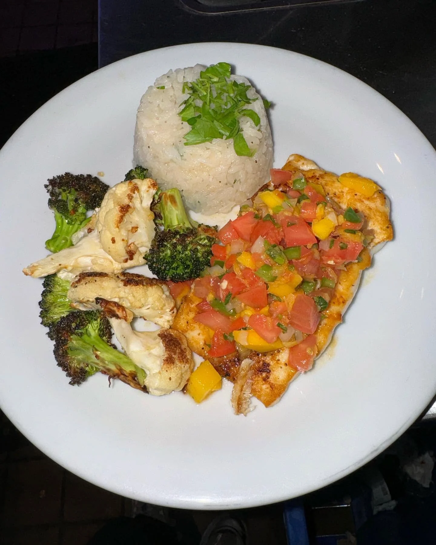 Plate of baked fish topped with diced tomatoes, green bell peppers, and yellow bell peppers, served with roasted broccoli and cauliflower, and a side of shaped white rice garnish with green herbs.