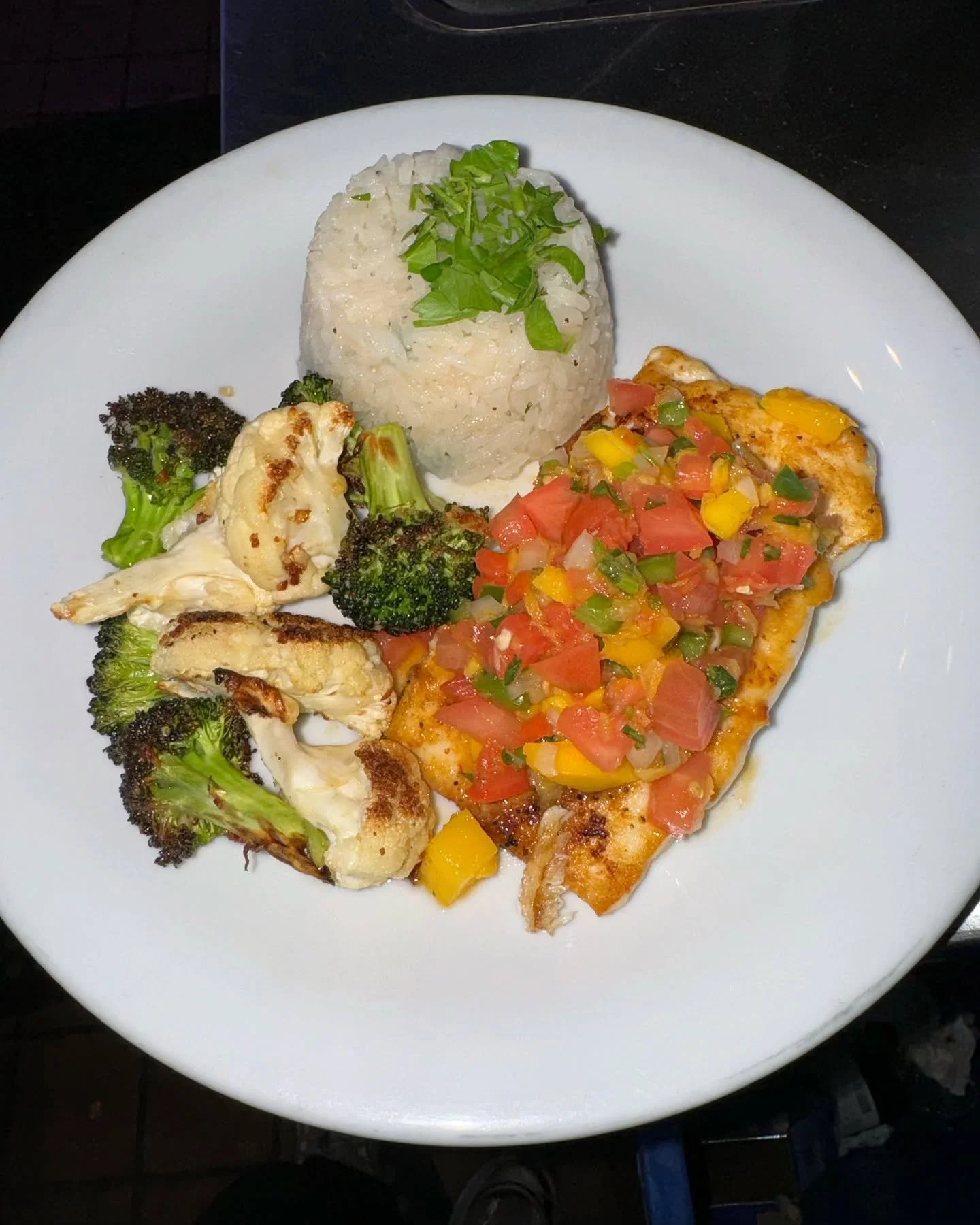 Plate with grilled cauliflower and broccoli, serving of rice topped with chopped green herbs, and a piece of fish topped with diced tomatoes, green peppers, and yellow peppers.