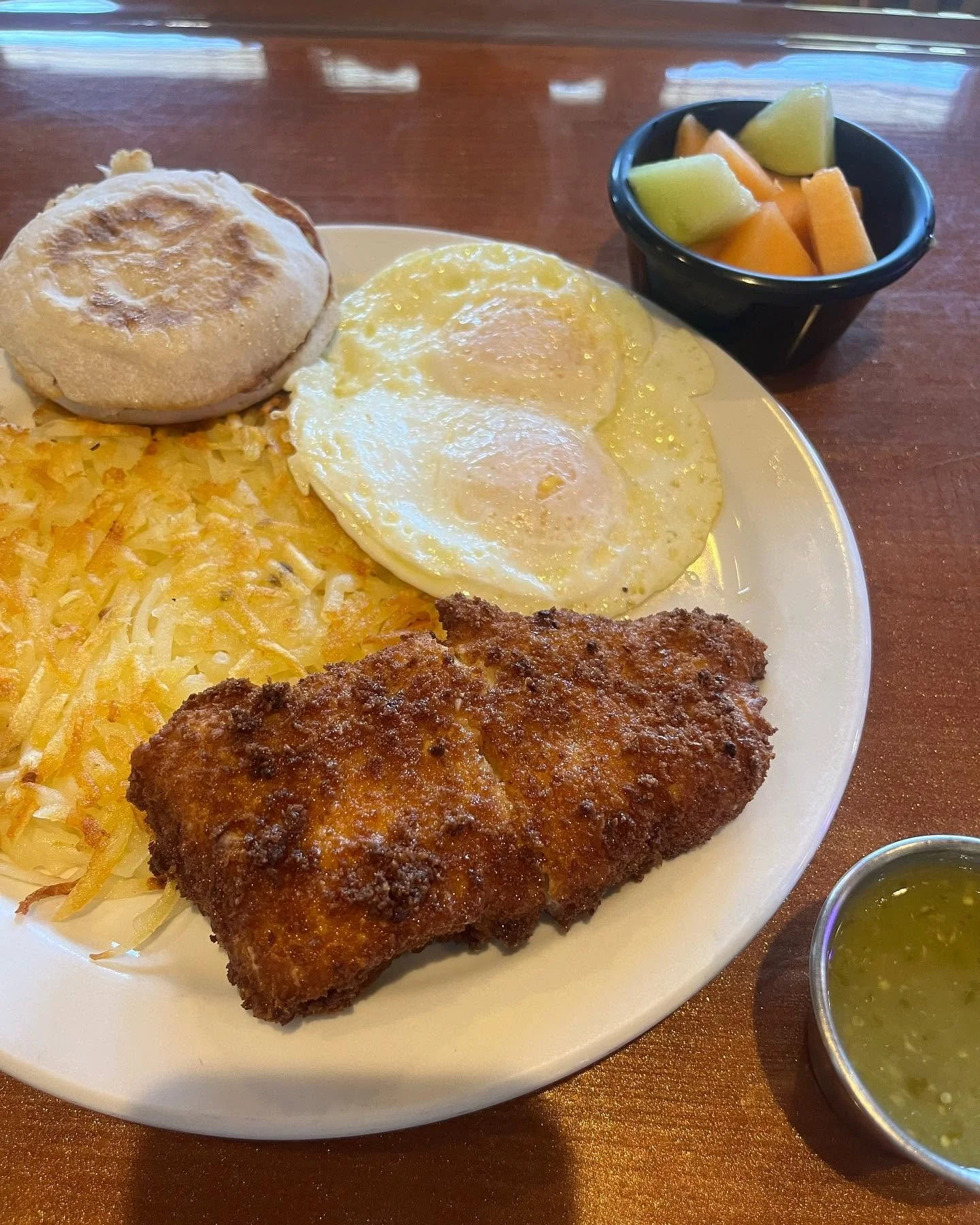 A plate of breakfast food including two sunny-side-up eggs, cheesy hash browns, a biscuit, and a breaded fried pork chop, with side bowls of fresh fruit and green sauce.