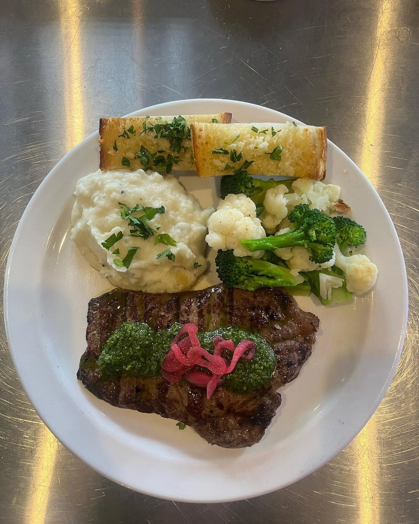 Plate of food with grilled steak topped with green sauce and pickled red onions, roasted cauliflower with green herbs, mashed potatoes with herbs, fried bread sticks garnished with herbs, and sautéed broccoli, on a white plate.