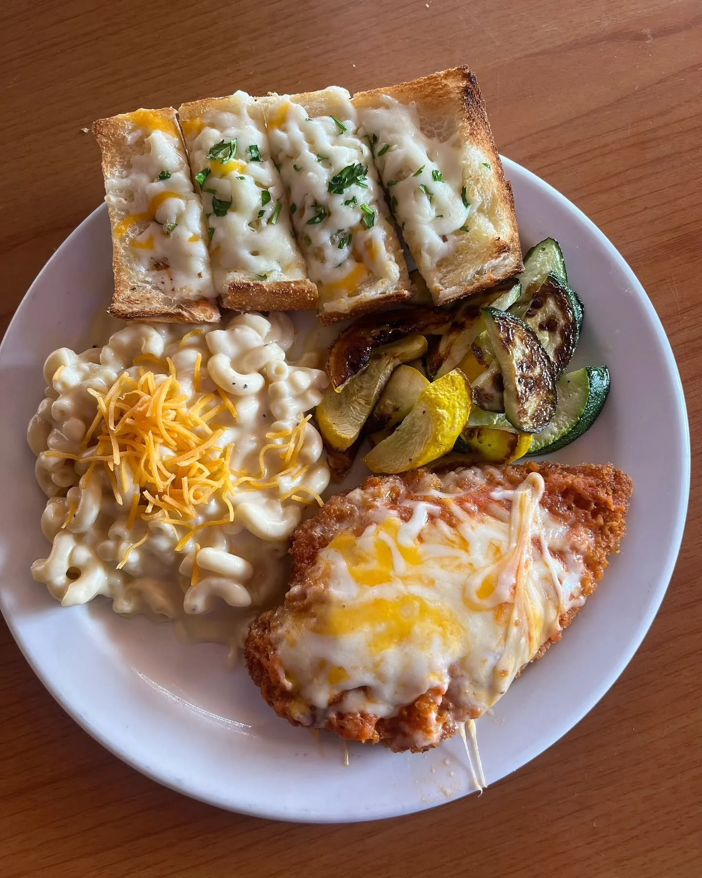 A white plate of breakfast food includes baked cheesy bread, macaroni and cheese, grilled zucchini and yellow squash, and a breaded chicken cutlet with melted cheese and topped with a fried egg.