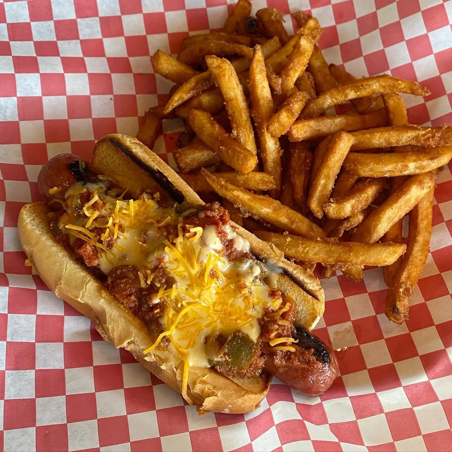 Hot dog with cheese, relish, and chili, served with a side of French fries on red and white checkered paper.