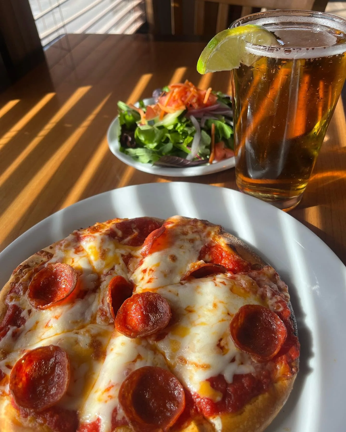 Close-up of a pepperoni pizza on a white plate, with a side salad in a small bowl, and a glass of iced tea with lime on a wooden table.