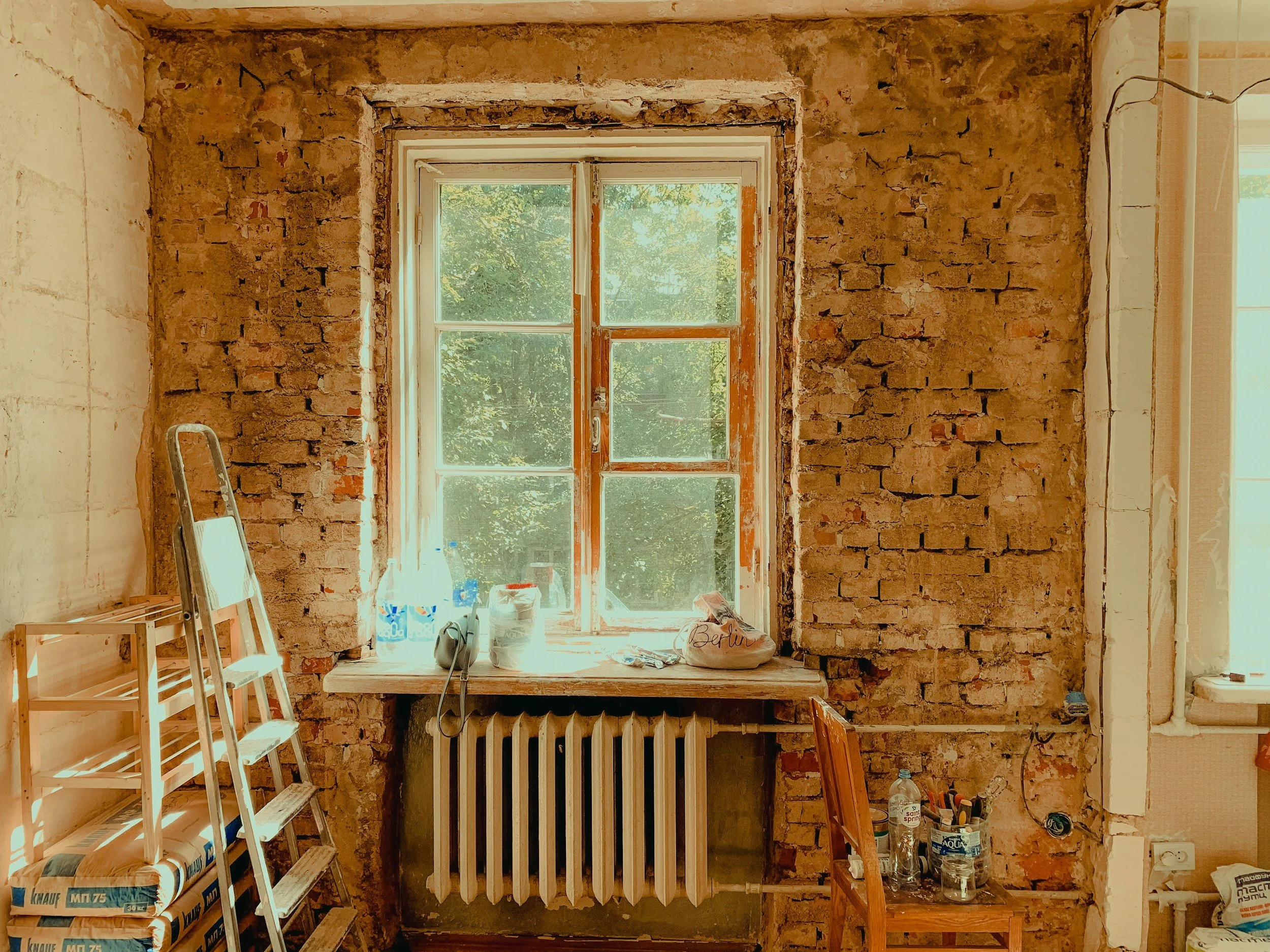 Room under renovation with exposed brick walls, a window showing greenery outside, construction supplies and tools, a ladder, and a small table with water bottles and painting supplies.