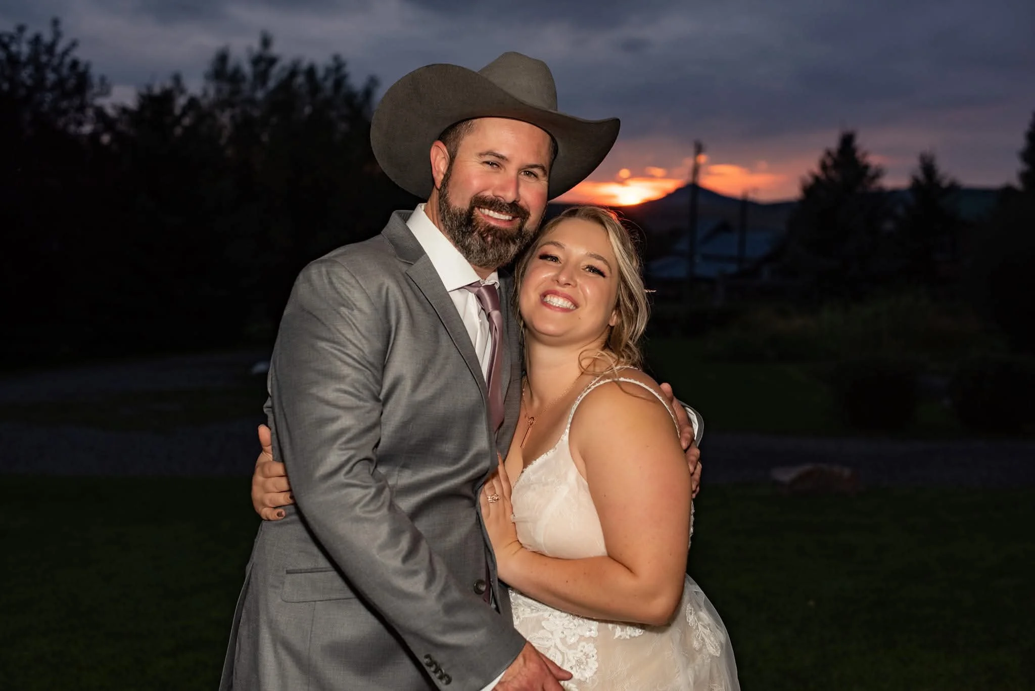 A couple dressed in wedding attire outdoors at sunset, with the groom wearing a gray suit and cowboy hat, and the bride in a white lace wedding dress, embracing and smiling.