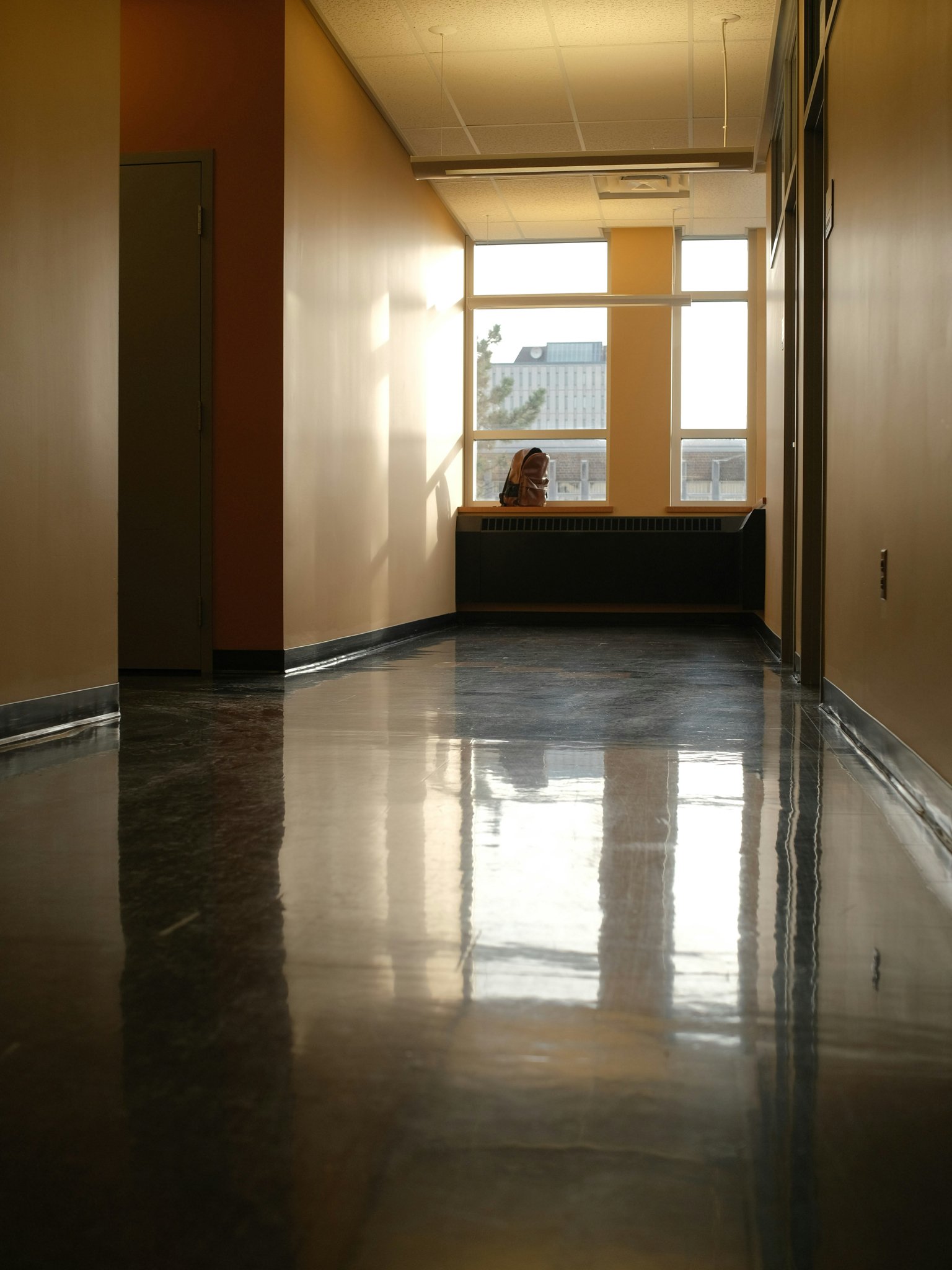 Empty hallway with reflective dark marble flooring, beige walls, and large windows at the end letting in natural light, with a bag on the window ledge.