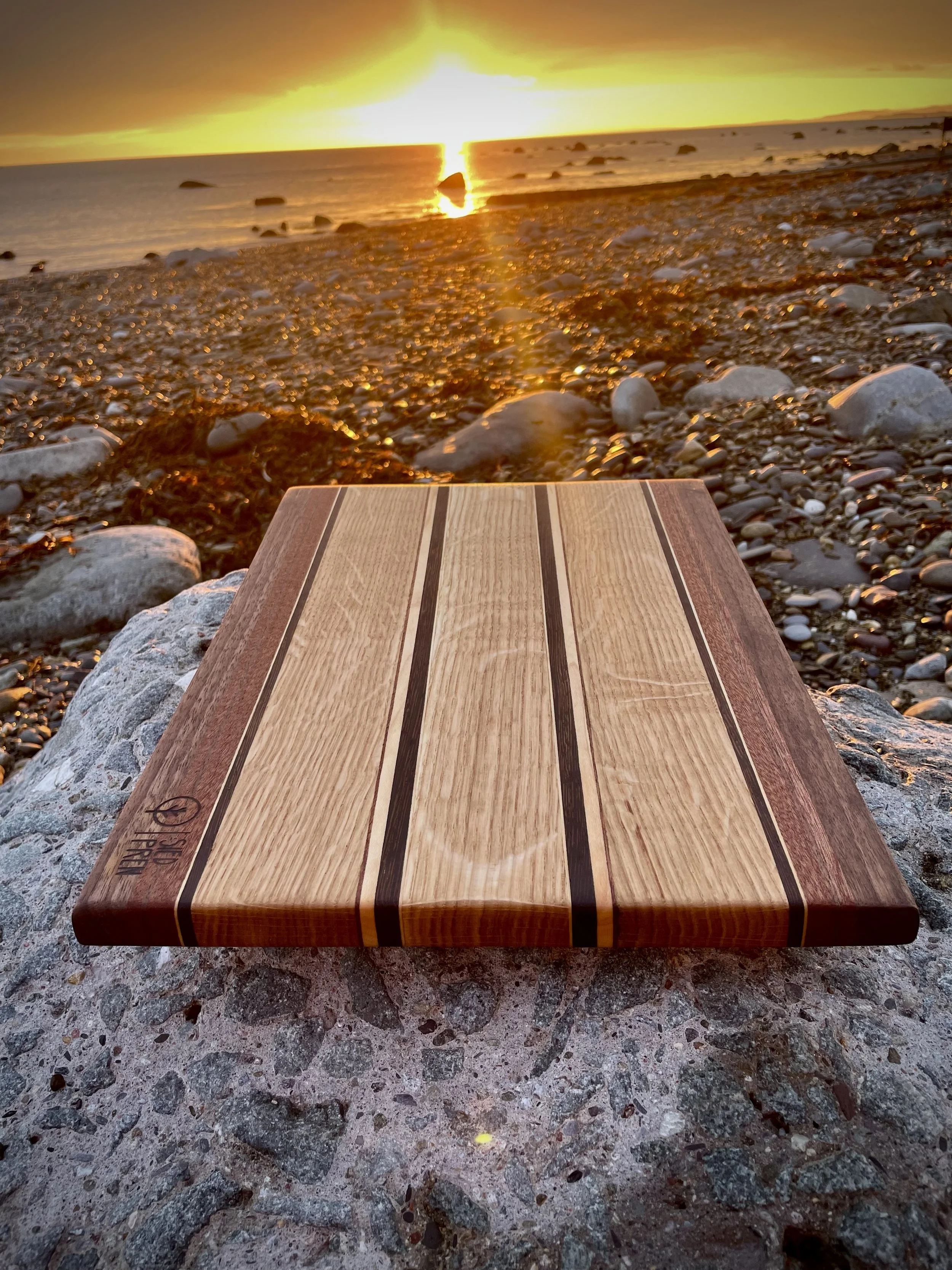 A wooden cutting board with decorative stripes on a rocky beach at sunset, with the ocean and a cloudy sky in the background.