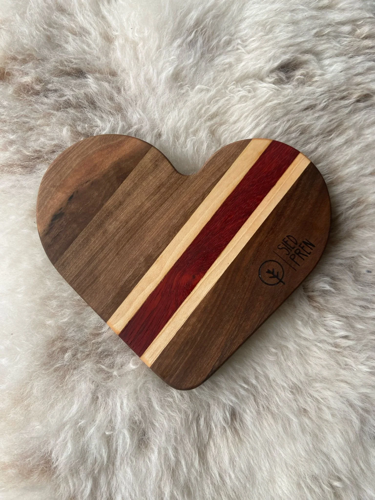A heart-shaped wooden cutting board with colored inlays of dark brown, light beige, and reddish-brown, resting on a soft, fluffy white surface.