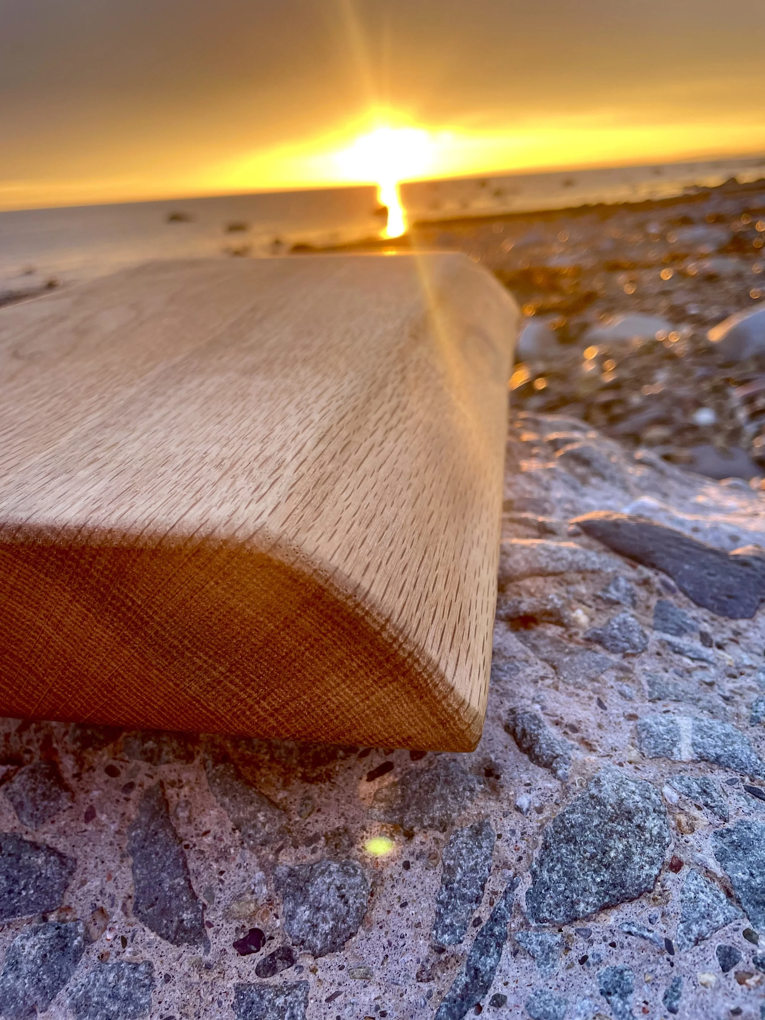 Close-up of a rock and pebble-covered beach with a wooden plank in the foreground, and a sunset over the ocean in the background.