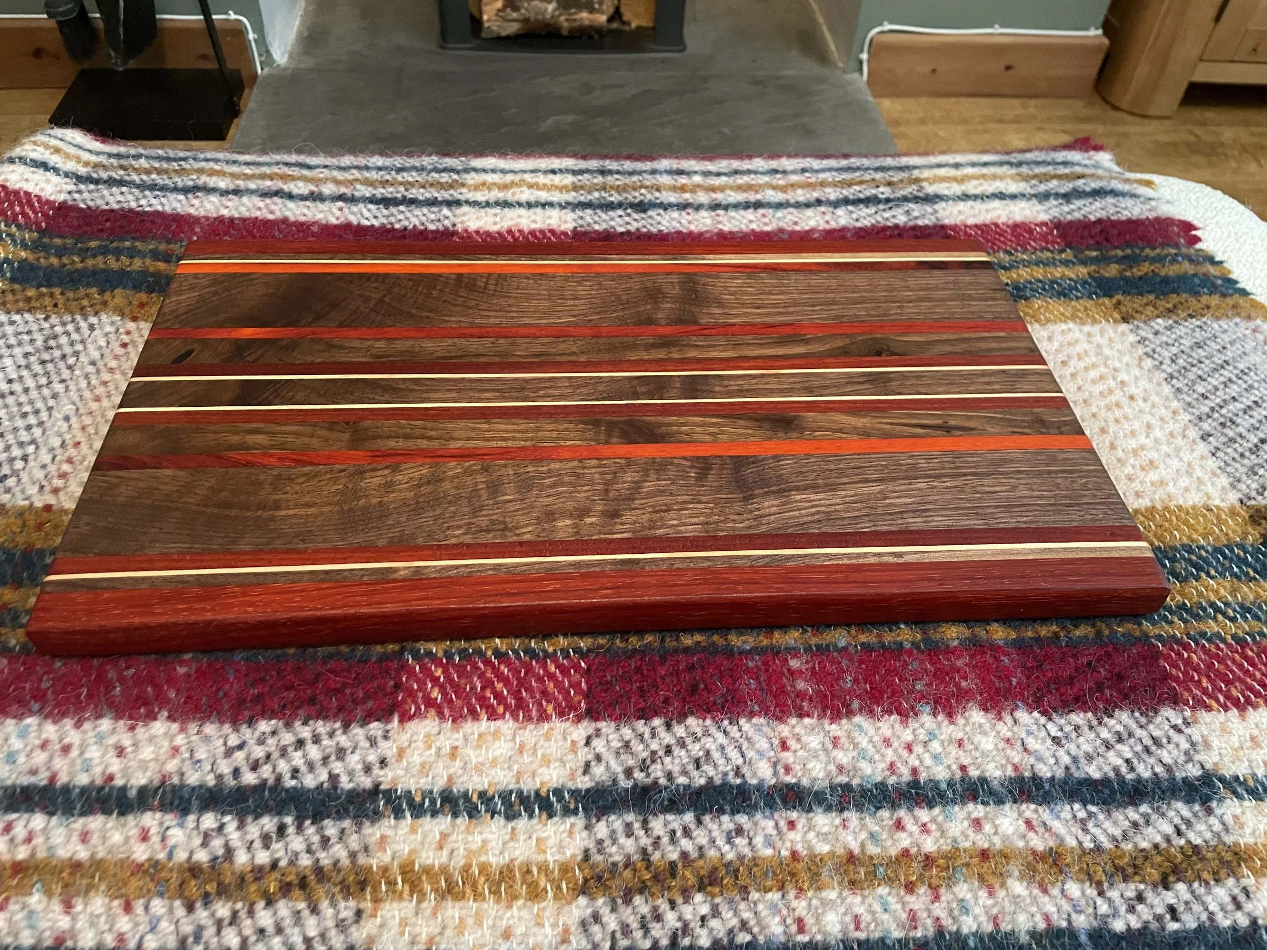 A wood cutting board with dark and reddish wood stripes on a multicolored woven carpet with red, white, and blue patterns. In the background, a fireplace with a stone hearth and a wood-log fire is visible.