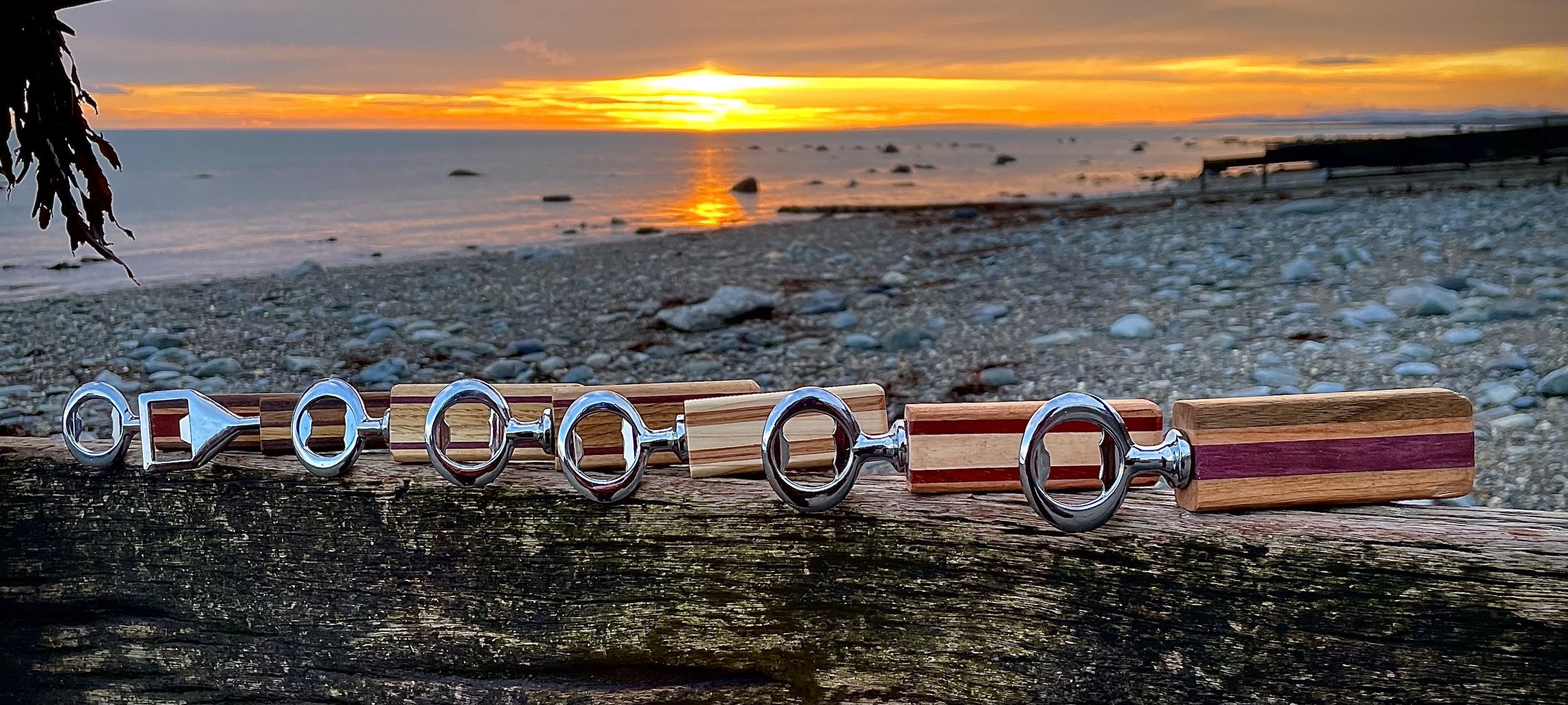 A row of wine corks with metallic holders on a weathered log, set against a beach and sunset background with rocks in the water.