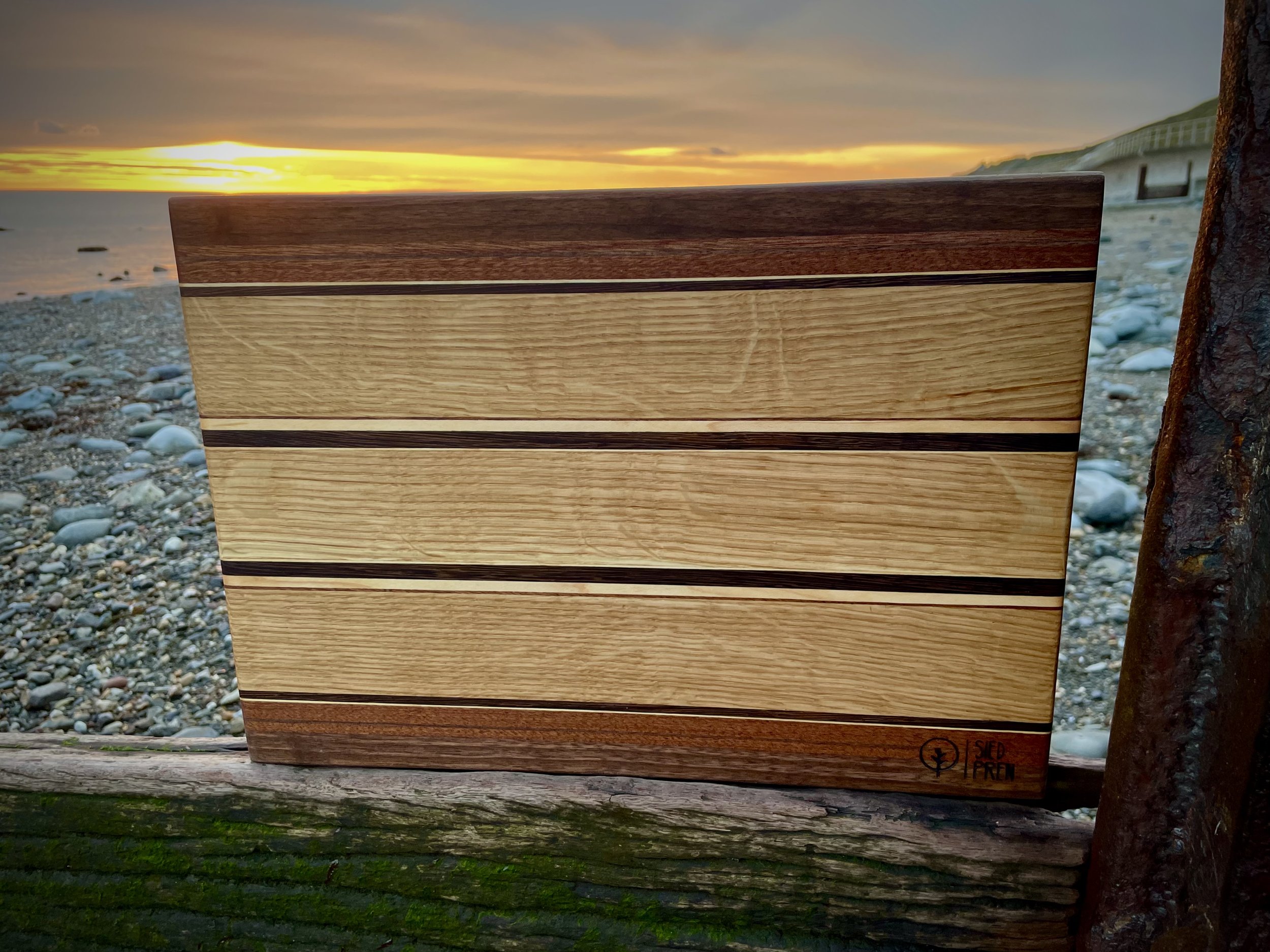 A close-up of a handcrafted wooden cutting board with alternating light and dark wood stripes, placed outdoors on a weathered wooden surface, with a rocky beach and sunset sky in the background.